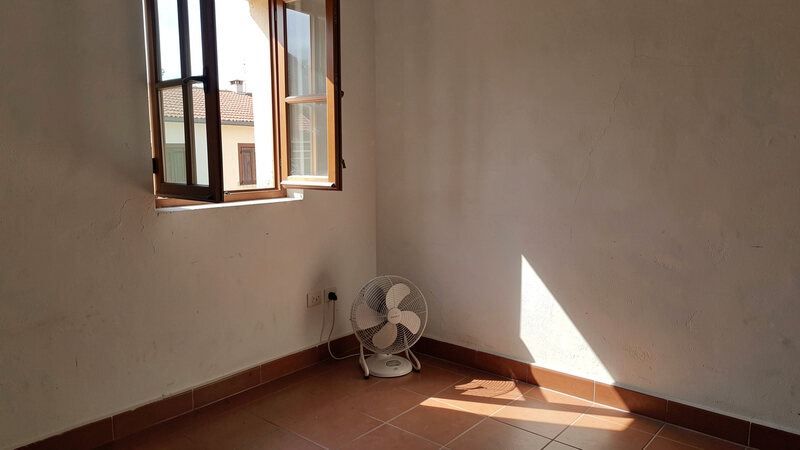 A room corner with an open window, a small fan placed low near the floor pointing towards the opening, and sunlight on the floor.